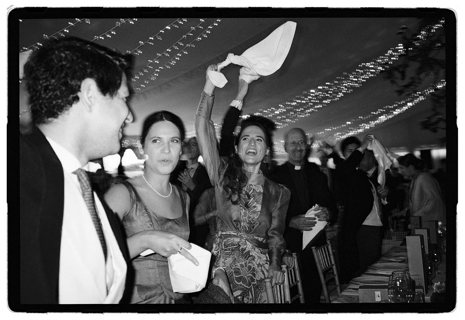 A woman at a formal event raising a napkin in celebration, surrounded by smiling people, including a man dressed as a priest and other guests, under string lights.