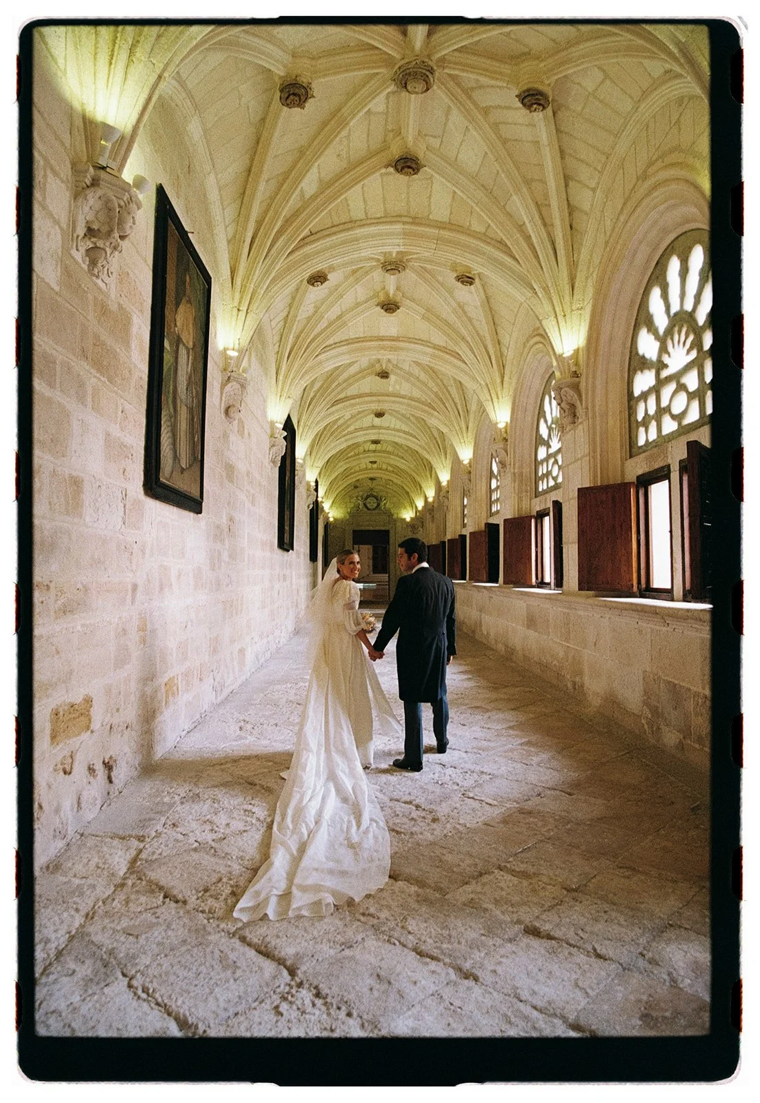 A bride and groom walking hand in hand in a historic stone hallway with vaulted ceilings and arched windows, holding hands and looking at each other.