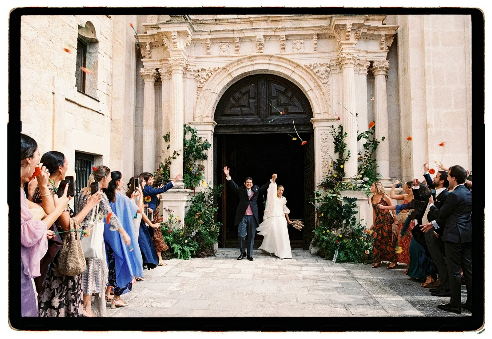 A newlywed couple exits a church, smiling and raising their hands, surrounded by friends and family throwing flower petals. The church has elaborate stone architecture with tall columns and ornate details. The crowd is dressed in formal attire, captu