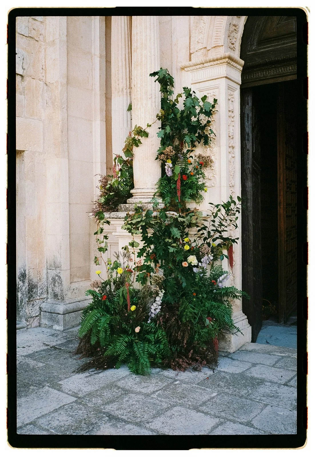 A flower arrangement with green foliage and colorful flowers placed outside a building with stone walls and classical architectural details.