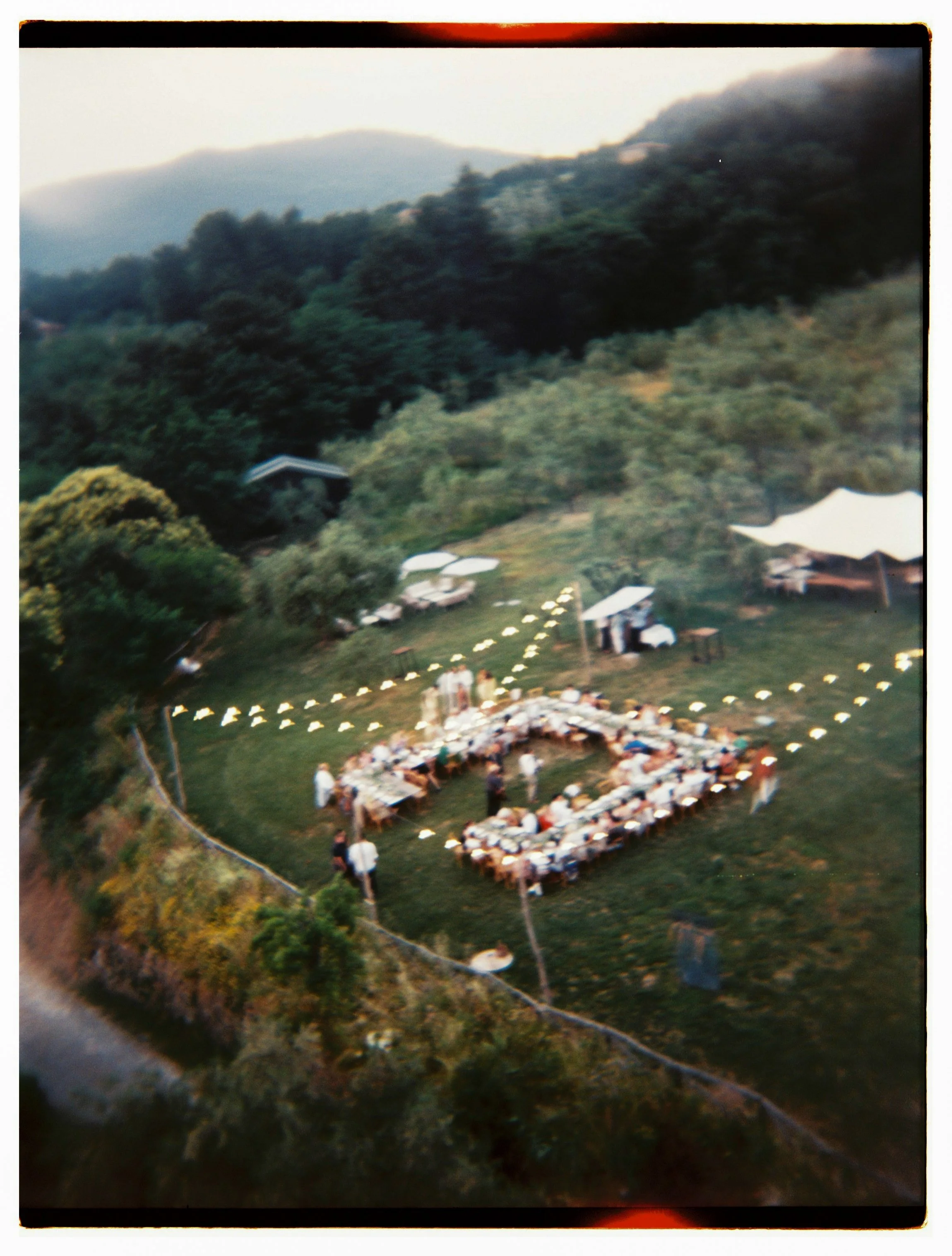 A large outdoor gathering in a U-shape formation with tables and chairs on a grassy area, surrounded by trees and hills, at sunset or dusk.
