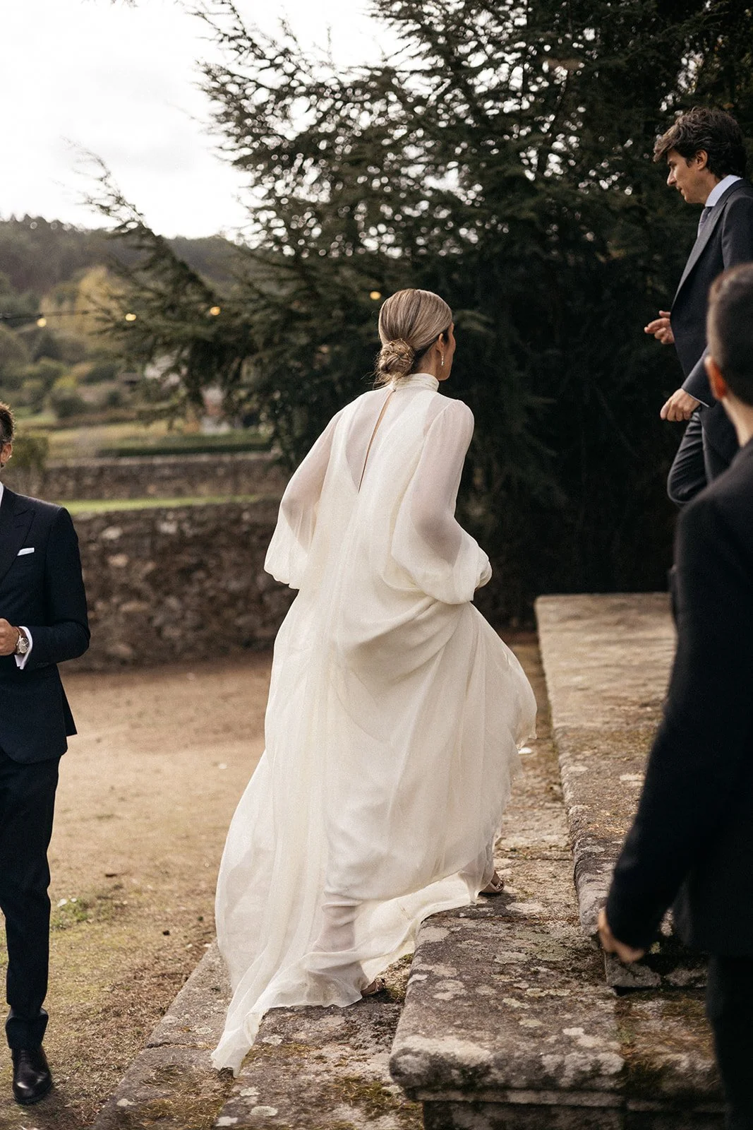A bride in a flowing white wedding dress descending stone steps outdoors, surrounded by formally dressed guests, under cloudy skies with a large tree in the background.