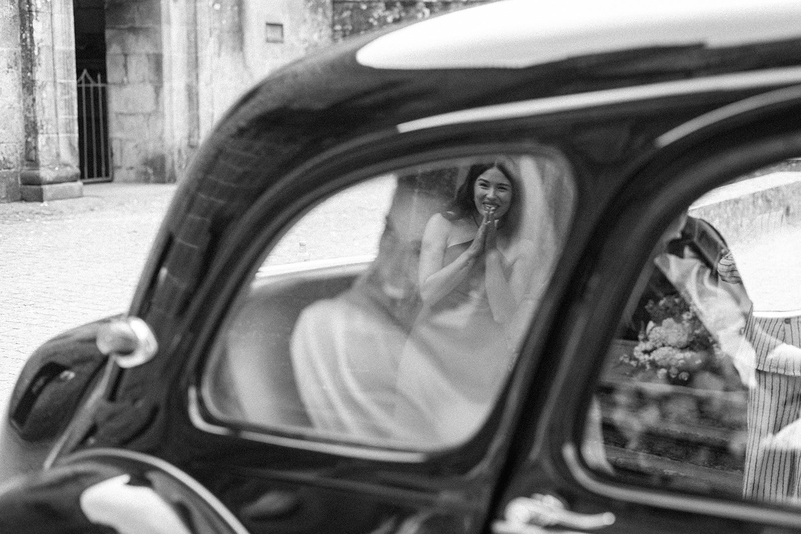 A woman smiling and clapping her hands, reflected in the window of a vintage car, with a person partially visible nearby and a street scene in the background.