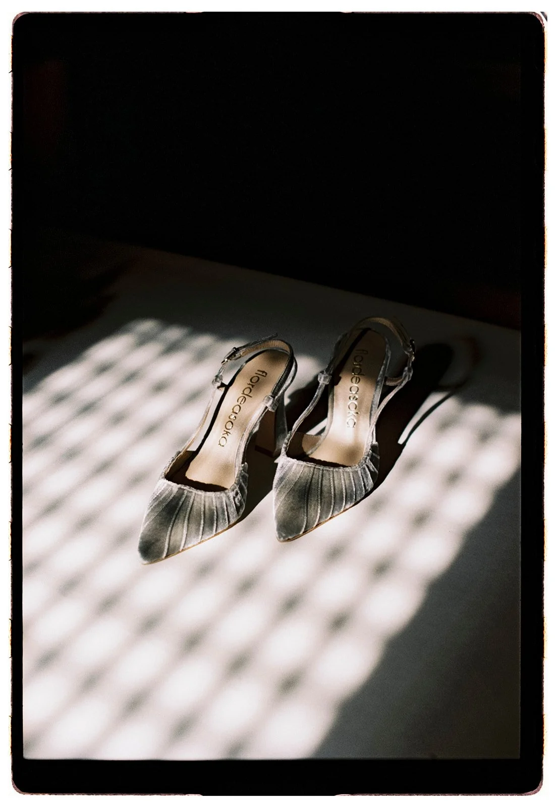 A pair of gray, pointed-toe high heels with a strap, placed on a surface with light and shadow pattern from window blinds.