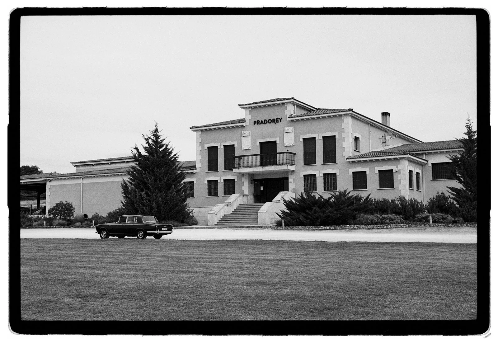 A black and white photo of a large, two-story building with a sign that reads "PRADOREY." There is a car parked in front of the building, and the scene includes some trees and a grassy field.