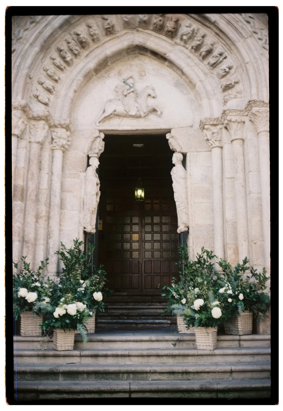 Stone church entrance with ornate archway, sculptures, and floral arrangements at the steps.
