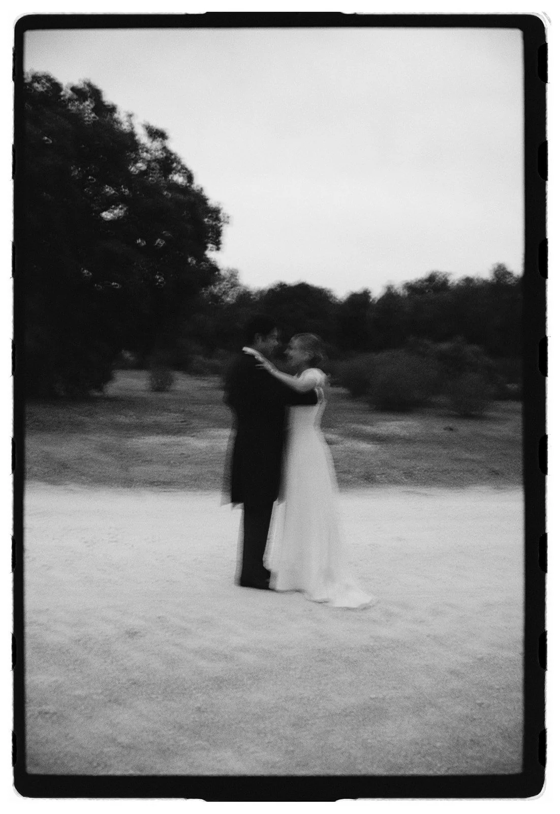 Black and white photo of a couple dancing outdoors, with trees in the background.