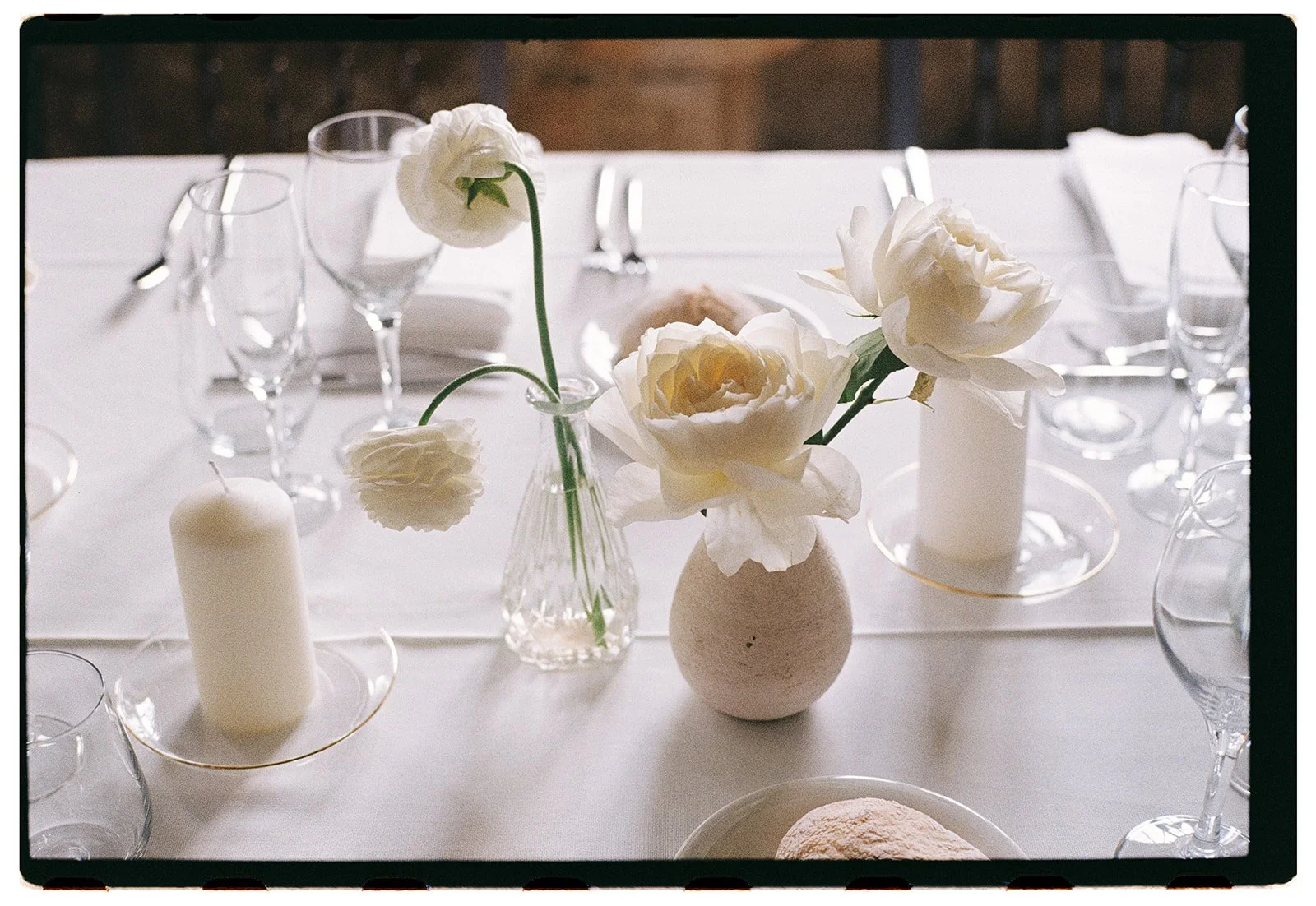 Elegant table setting with white flowers in a textured vase, surrounded by candles, glassware, and a white tablecloth.