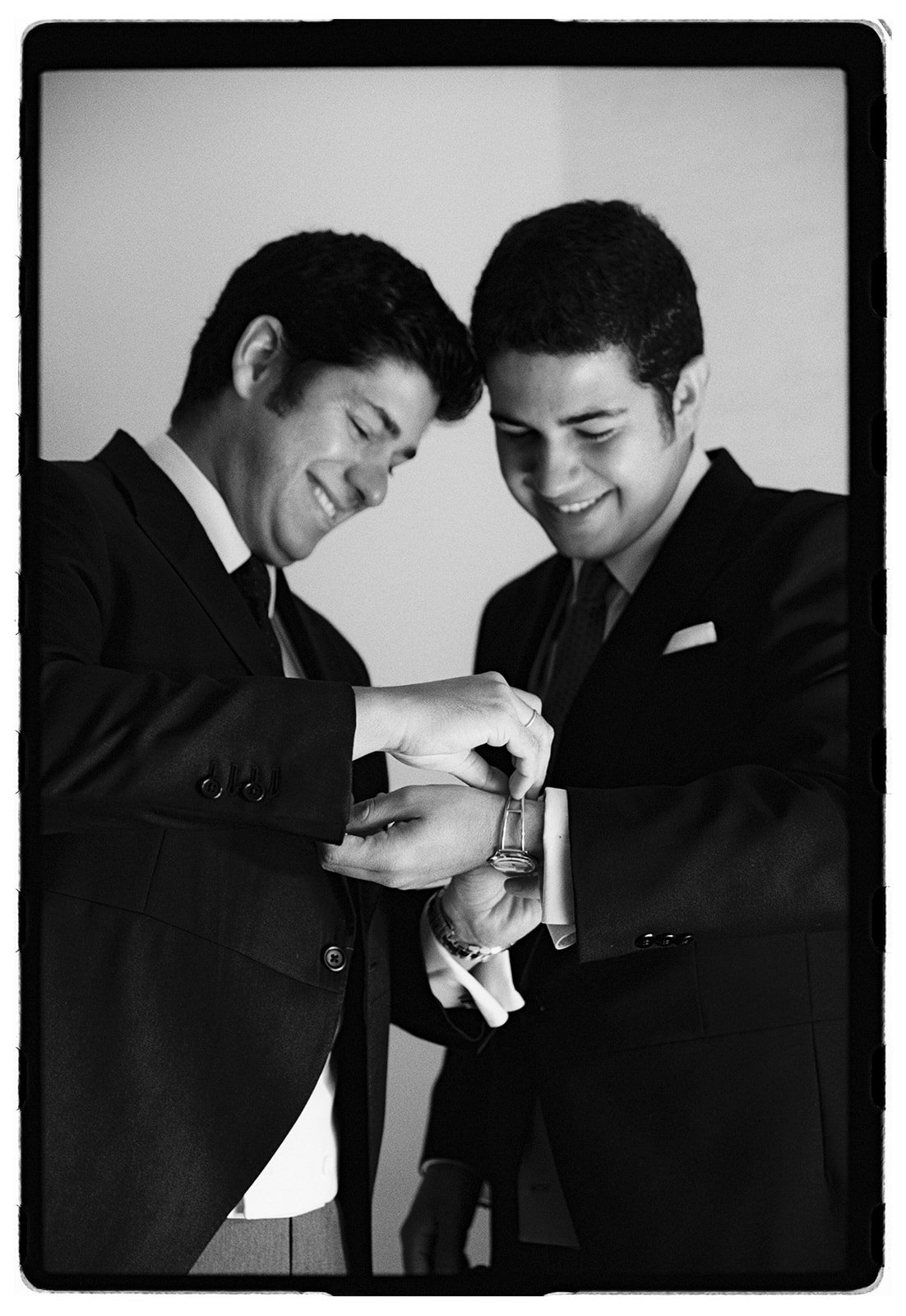 Two men in suits smile as one helps the other put on a wristwatch.