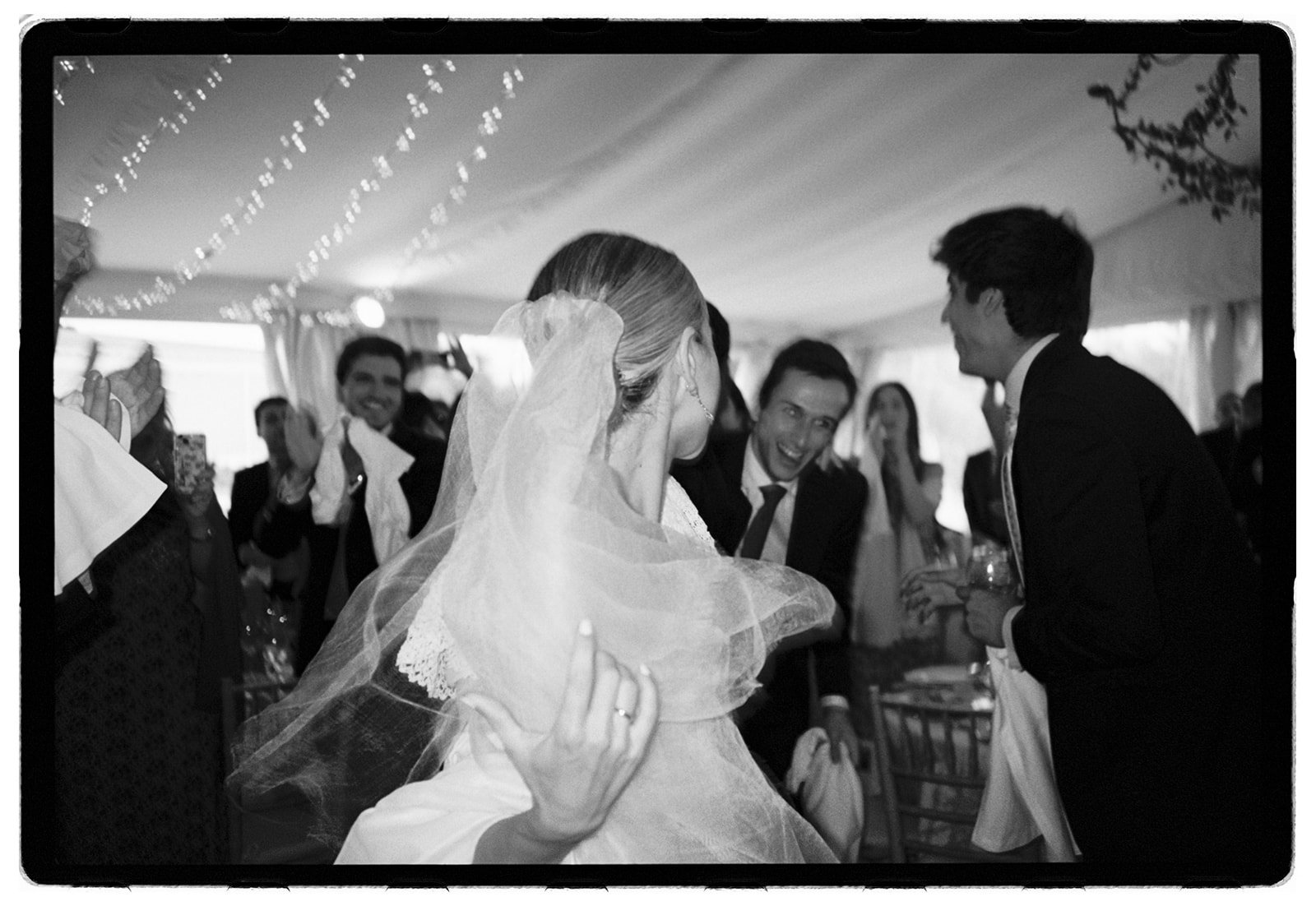 Black and white photo of a wedding reception. A bride with a veil is dancing and smiling, surrounded by guests who are clapping and cheering.