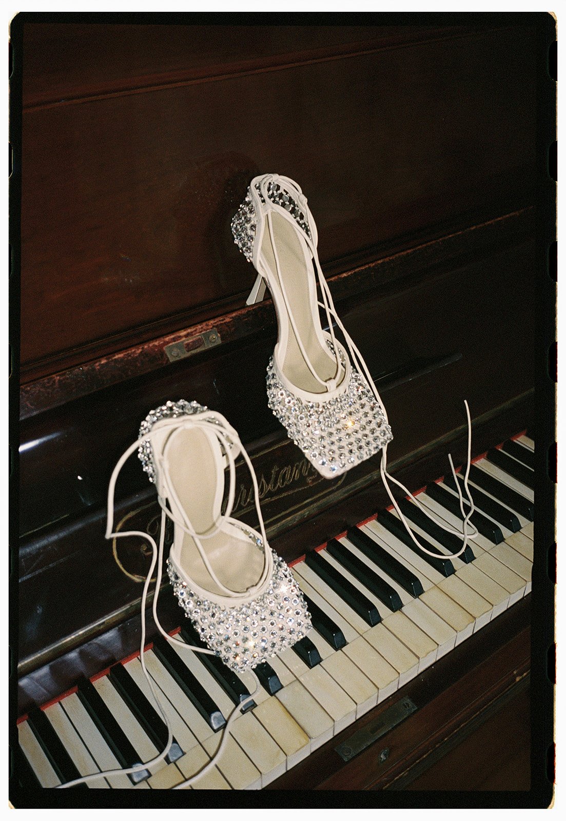 A pair of ornate, rhinestone decorated high-heeled shoes resting on piano keys. Bottega Veneta