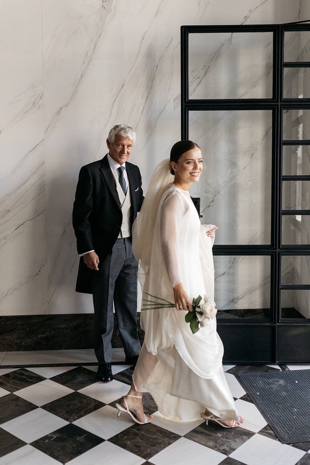 A bride in a white wedding dress and veil holding a small bouquet walks past an older man in a black tuxedo with striped pants, in a modern indoor setting with marble walls and a black and white checkered floor.