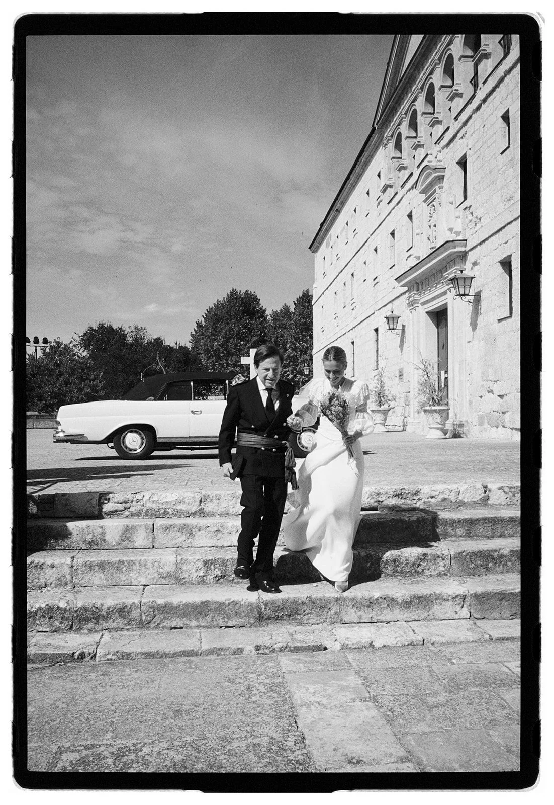 A black and white photo of a bride in a white wedding dress holding a bouquet, being assisted by a man in a formal dark suit, as they descend stone steps outside a large stone building, with a vintage car parked nearby.