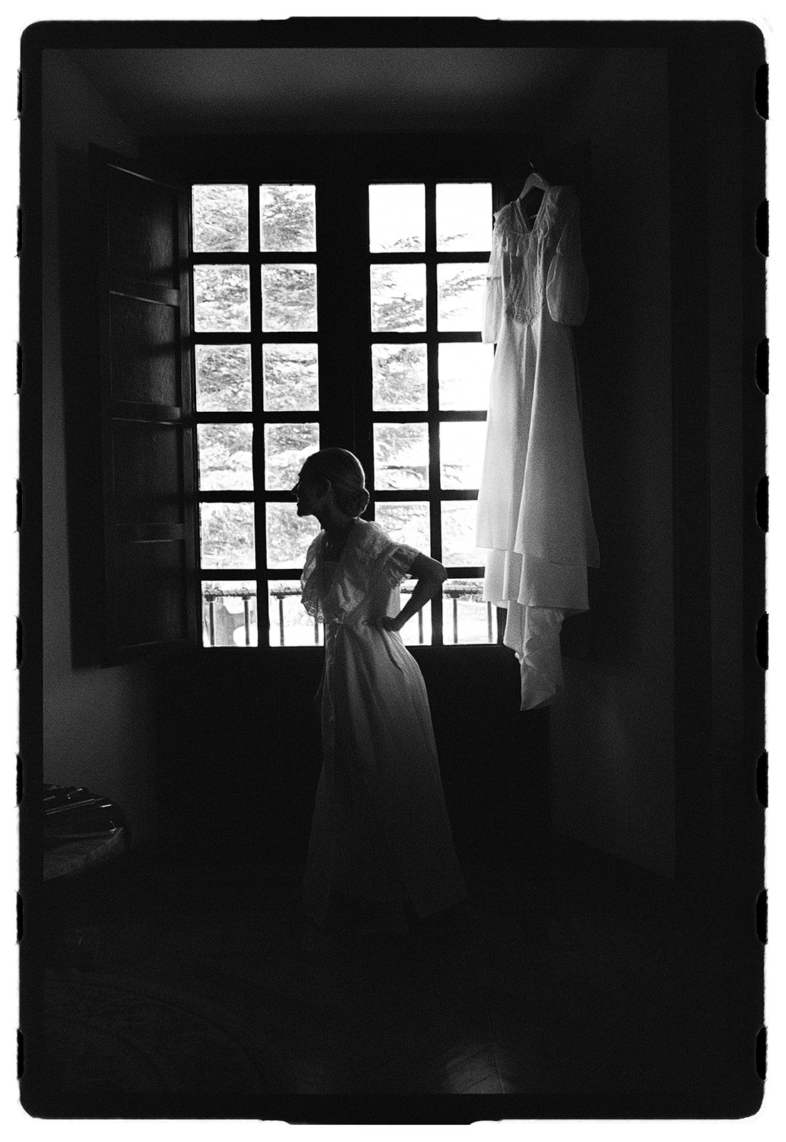 A silhouette of a woman in a dress standing in front of a large window with a wedding dress hanging beside her.