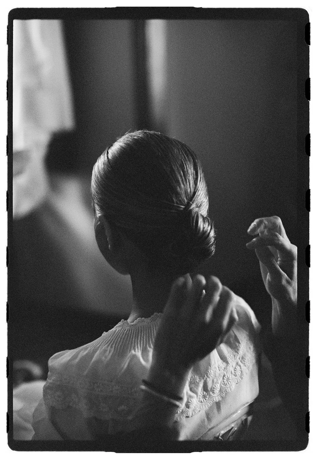 A woman with neatly styled hair is sitting, with another person adjusting her hair. The photo is in black and white.