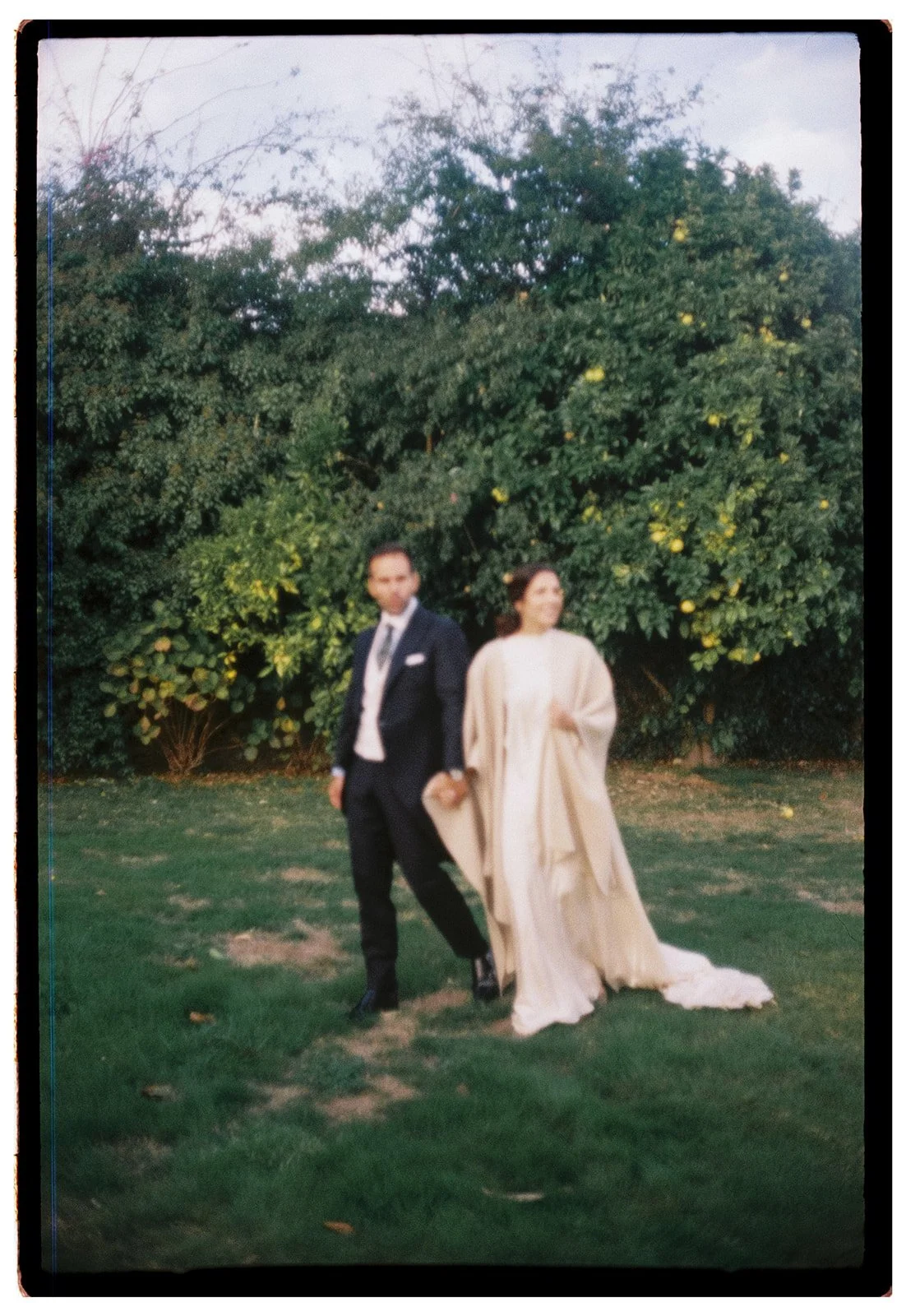 A bride and groom walking outdoors on grass with lush green bushes and yellow flowering trees in the background.