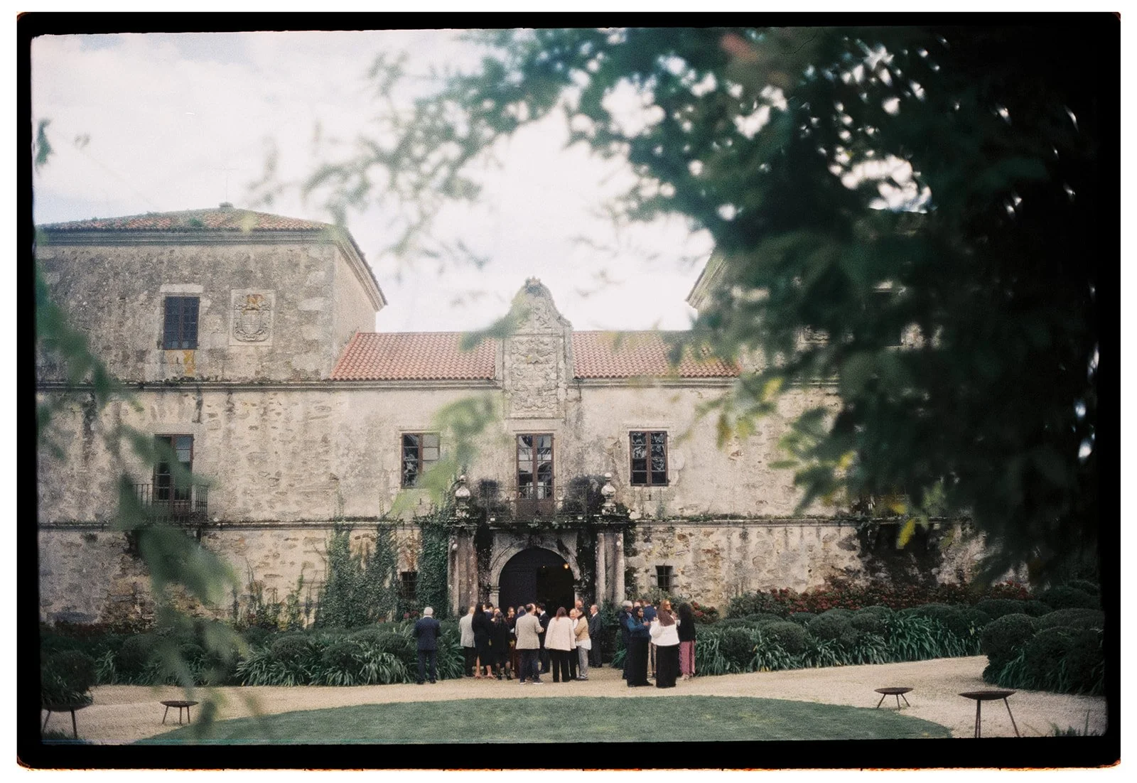 A group of people gathered outside an old stone castle with red tile roof, surrounded by greenery and trees, during daylight.