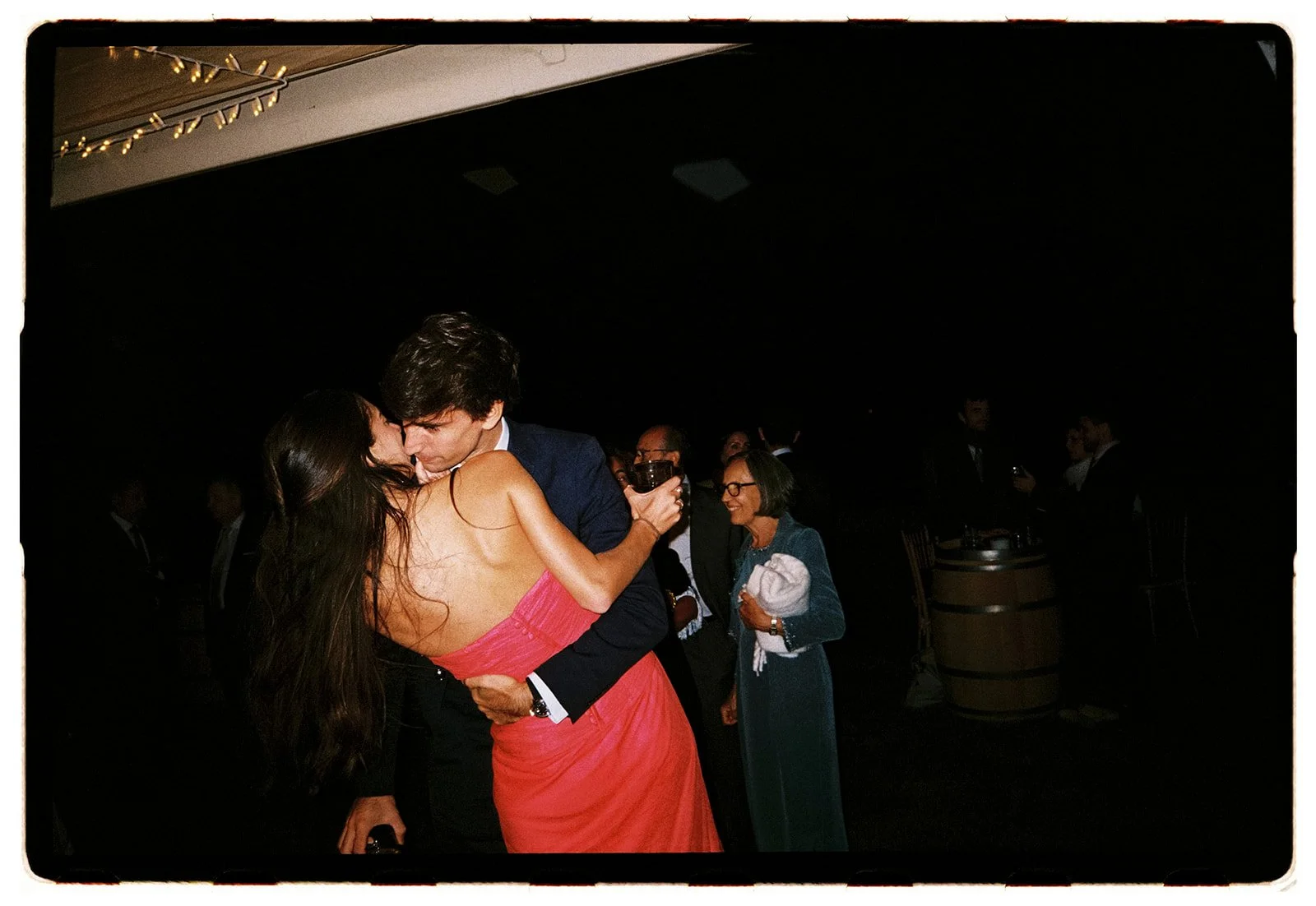 A couple dancing closely, the woman in a pink dress and the man in a suit, at a dimly lit social event, with other guests in the background holding drinks.