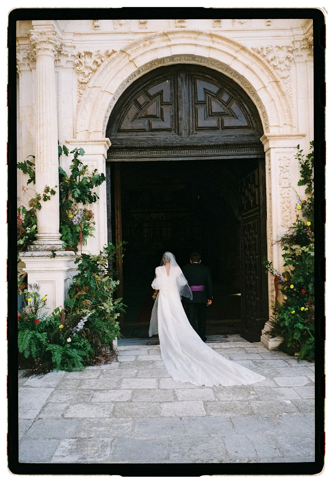 A bride in a white wedding gown and veil walking with a groom in a dark suit with a purple sash into a church entrance decorated with flowers.