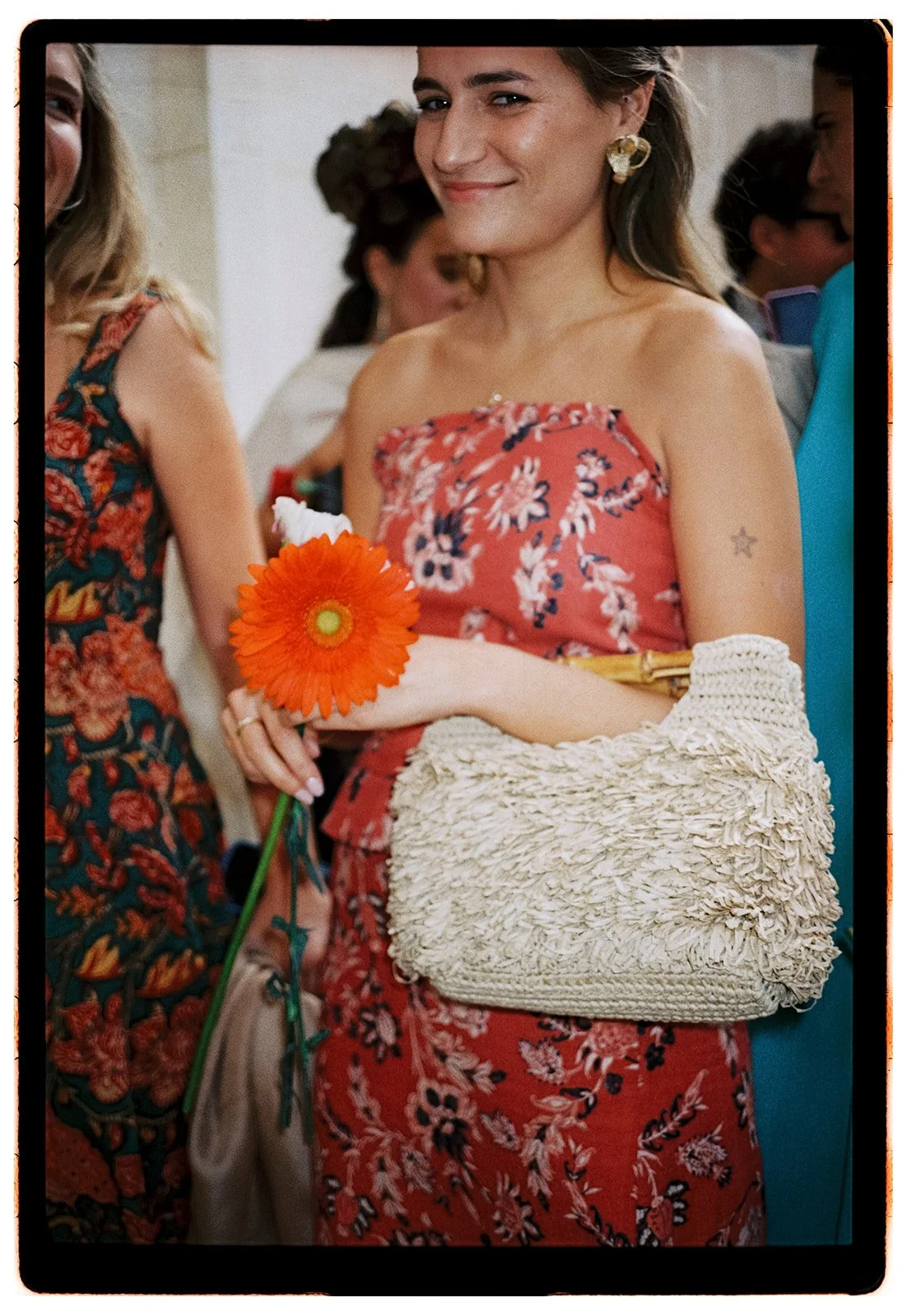 A woman in a red floral dress holding a bright orange flower, with a textured cream-colored bag over her arm and wearing star-shaped earrings.
