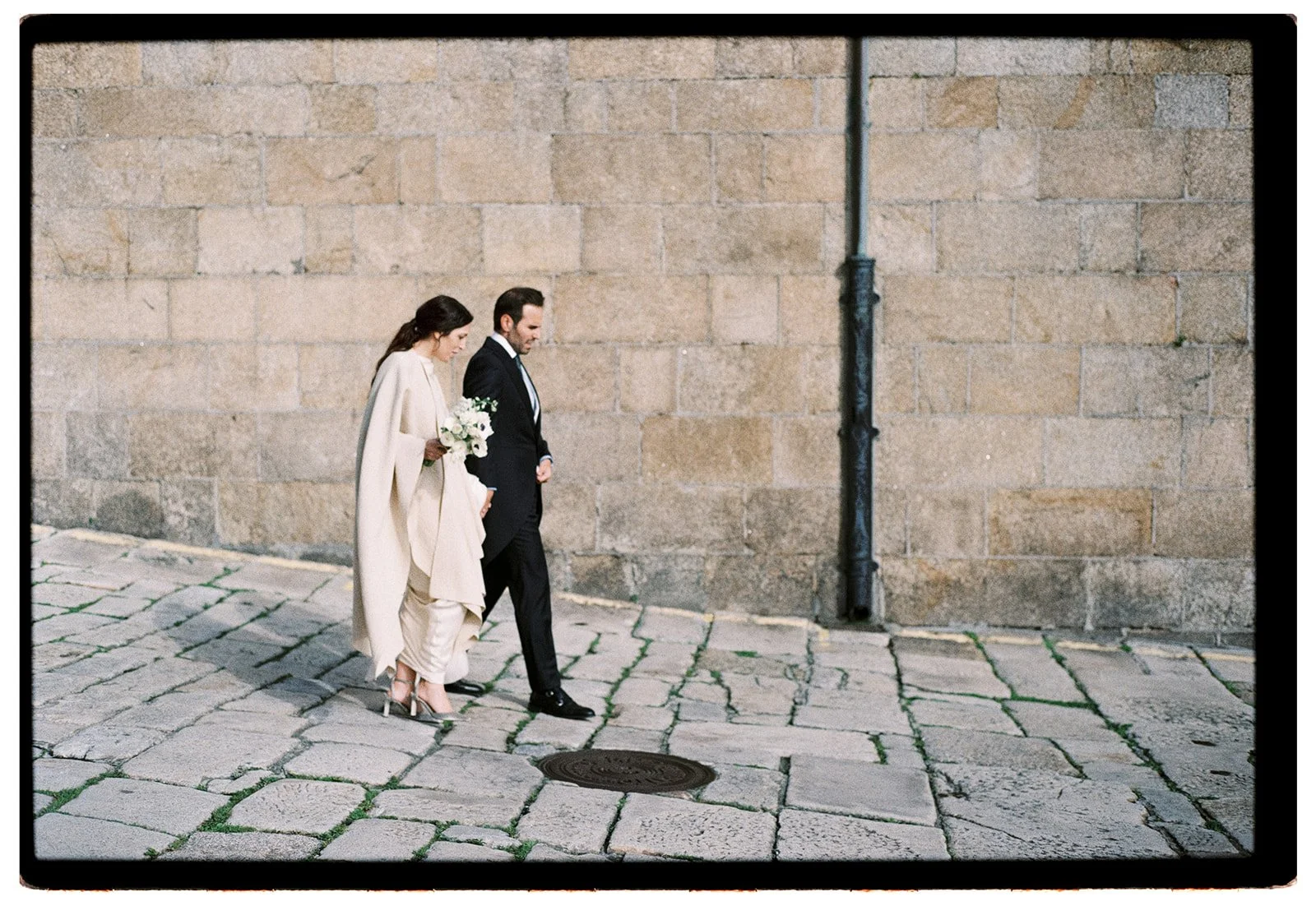 A bride and groom walking together on a cobblestone street next to a stone wall. The bride is holding a bouquet and wearing a cream-colored outfit, while the groom is in a black suit. Both are looking down as they walk.