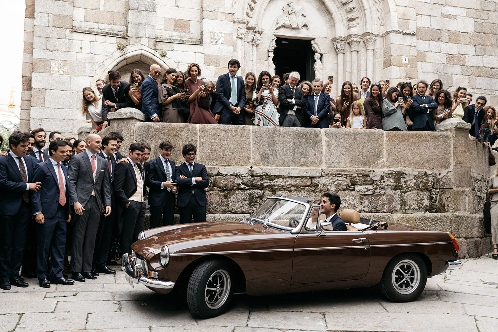 A man driving a vintage convertible car with beige interior, surrounded by a large group of people, many dressed in formal attire, at what appears to be a celebration or event outside an old stone building with an arched door.