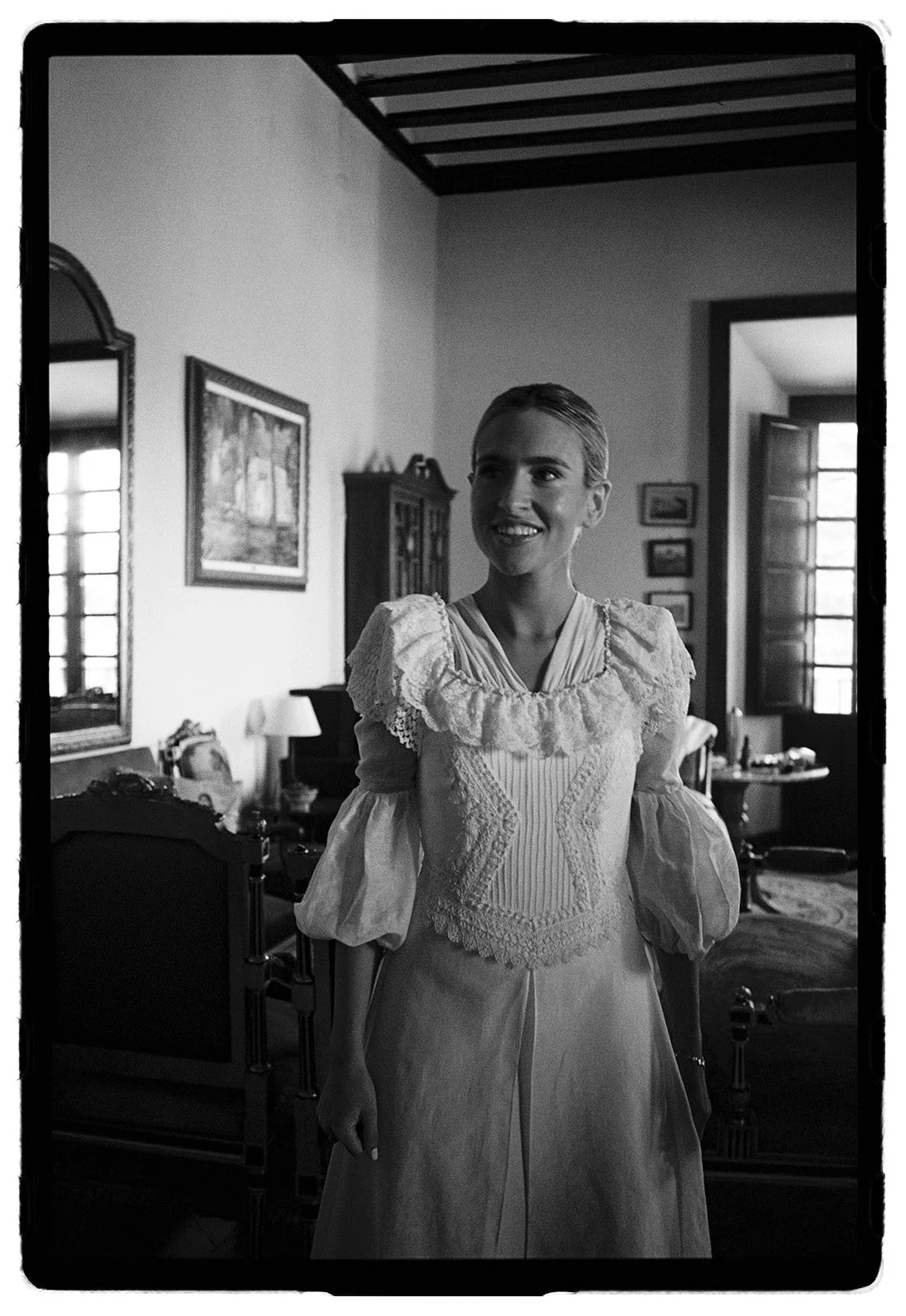 A woman in vintage dress standing in a spacious, well-lit room with antique furniture, cabinets, paintings, and open windows.