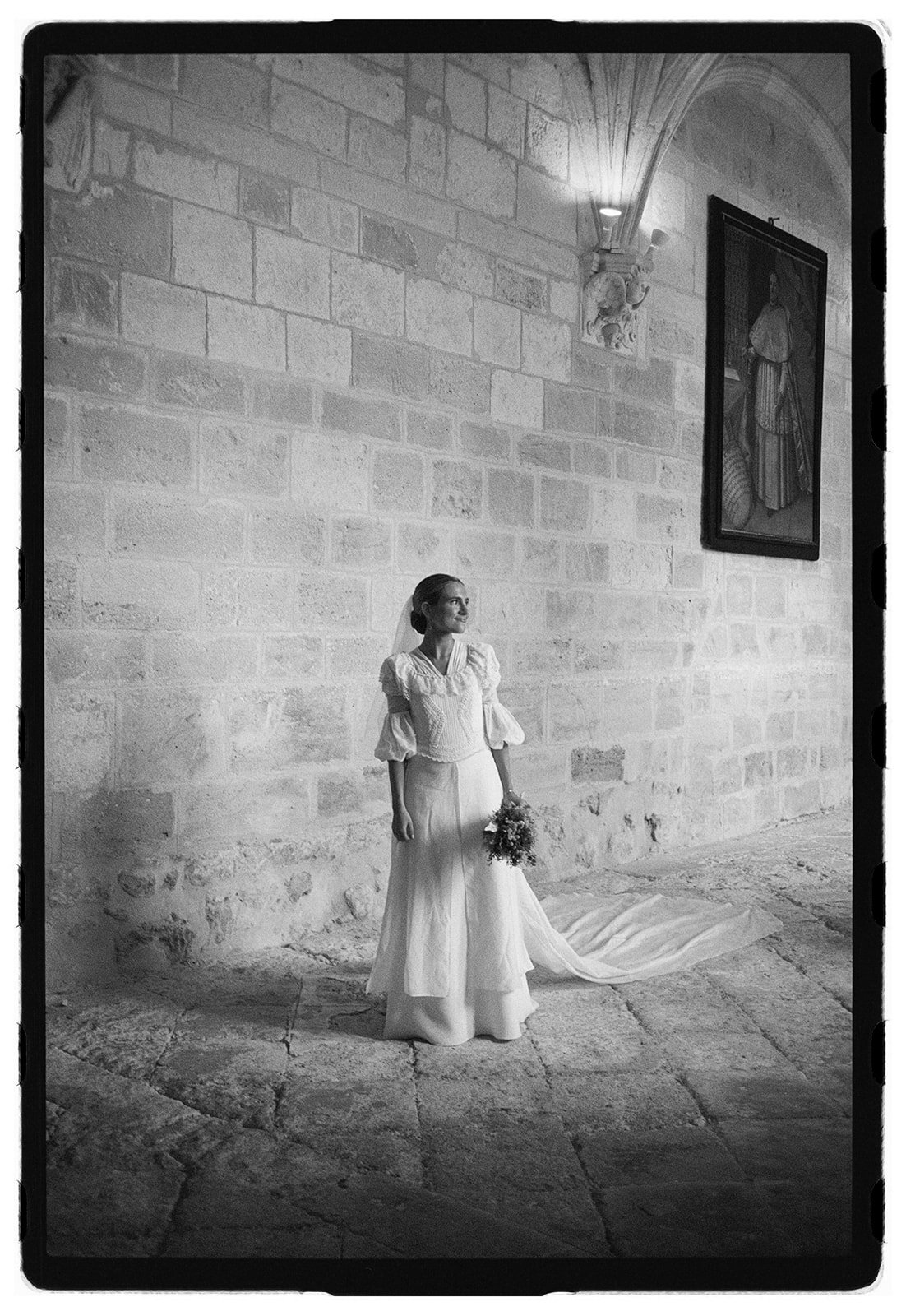A bride in a wedding dress holding a bouquet of flowers, standing inside a stone-walled church.