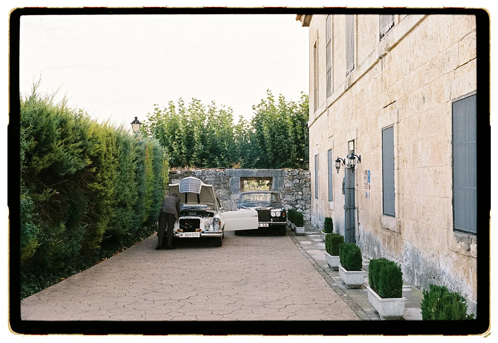 A person working on the engine of a vintage white car parked in a courtyard with greenery and stone walls.