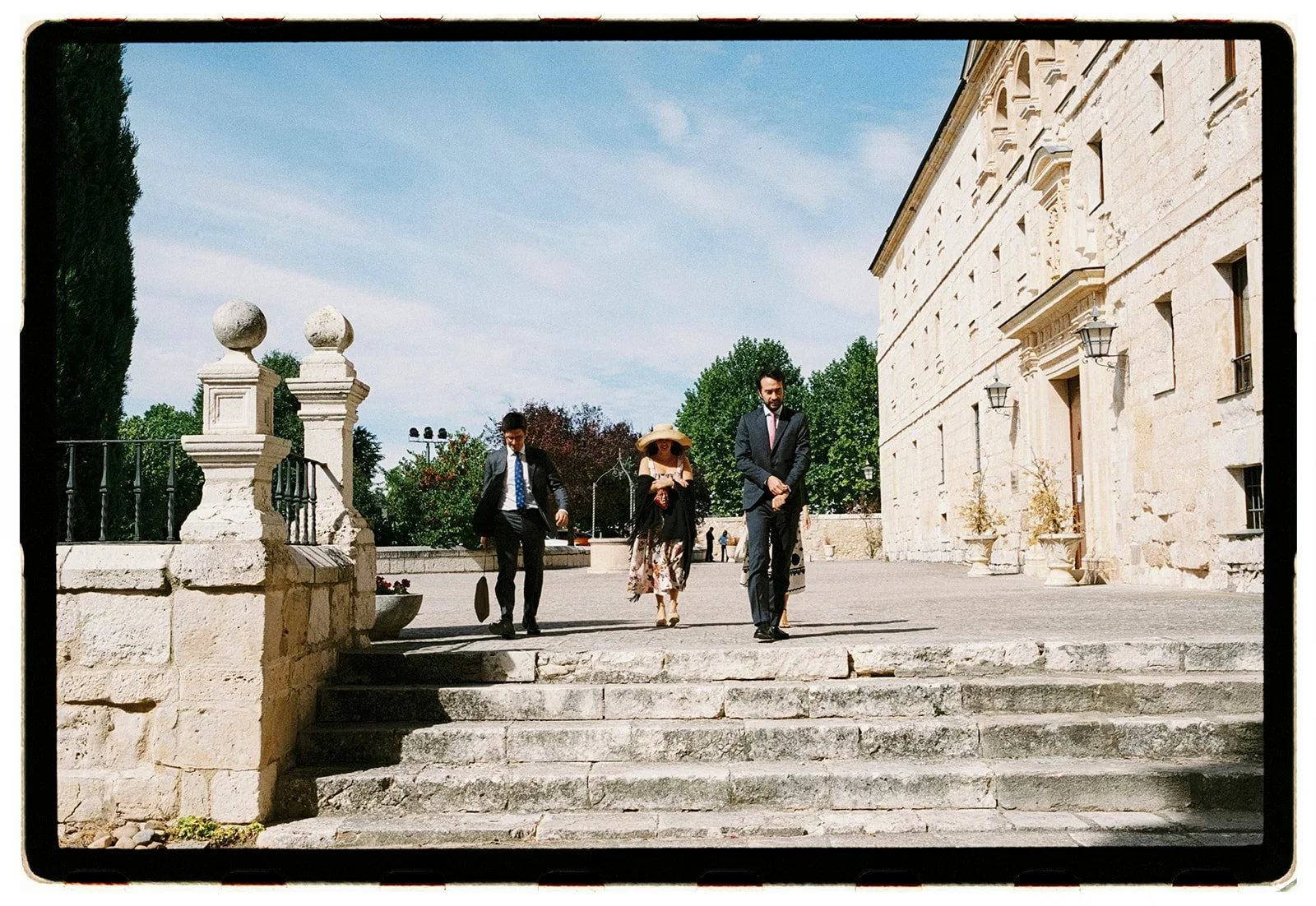 Group of three people walking down stone steps outside historic stone building on sunny day.