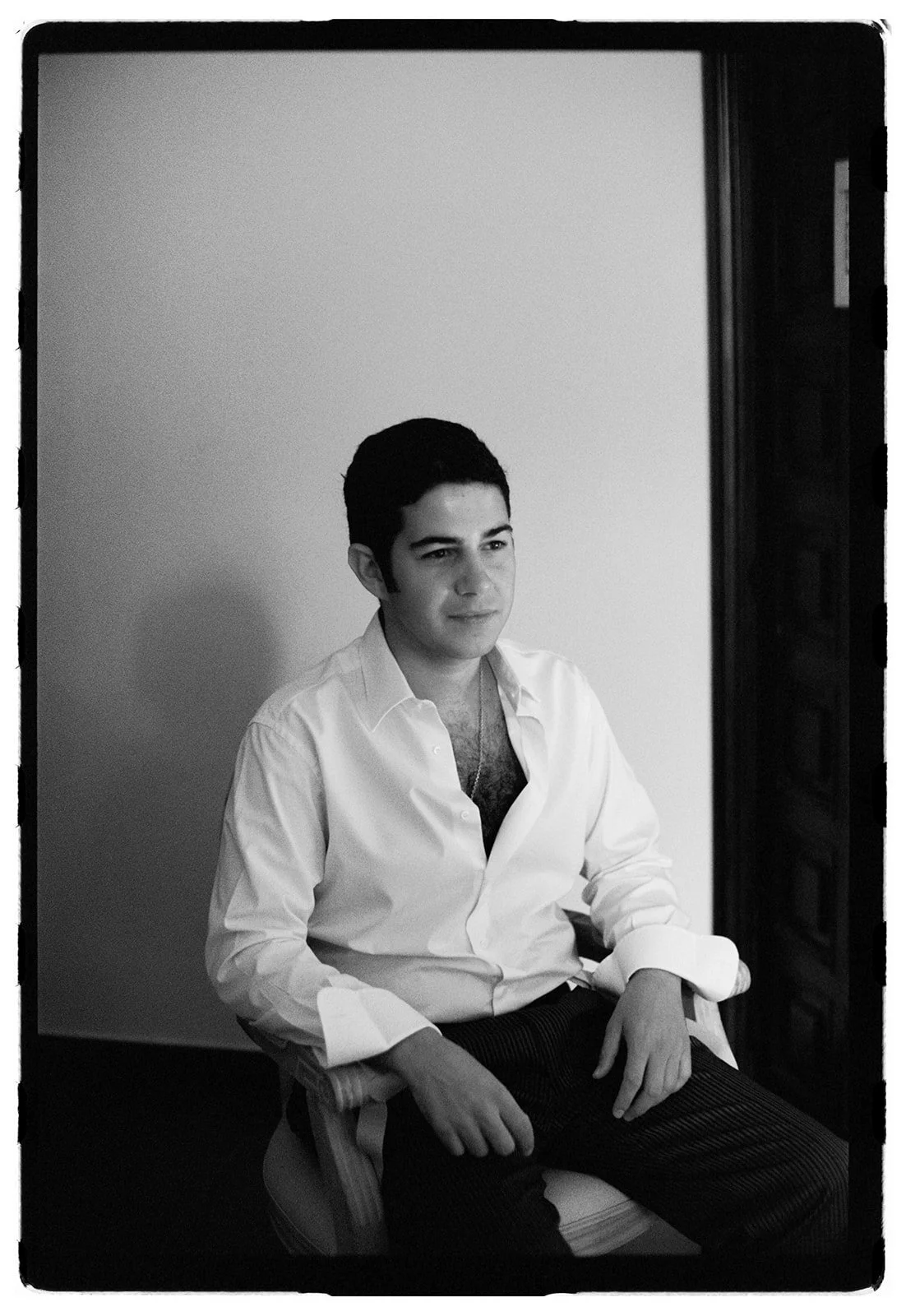 A young man with dark hair, wearing a partially open white dress shirt, sitting in a chair, gazing thoughtfully to the side.