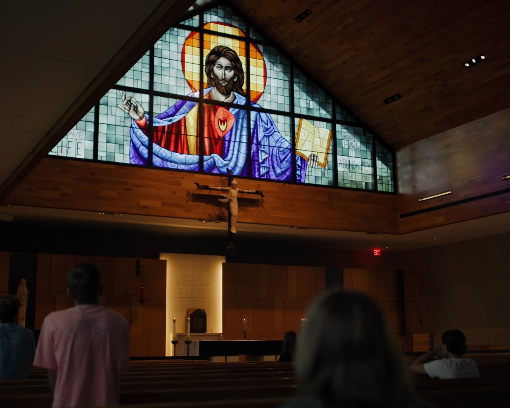 Interior of a church with a large stained glass window depicting a religious figure holding a book, a crucifix below, and people seated in pews.