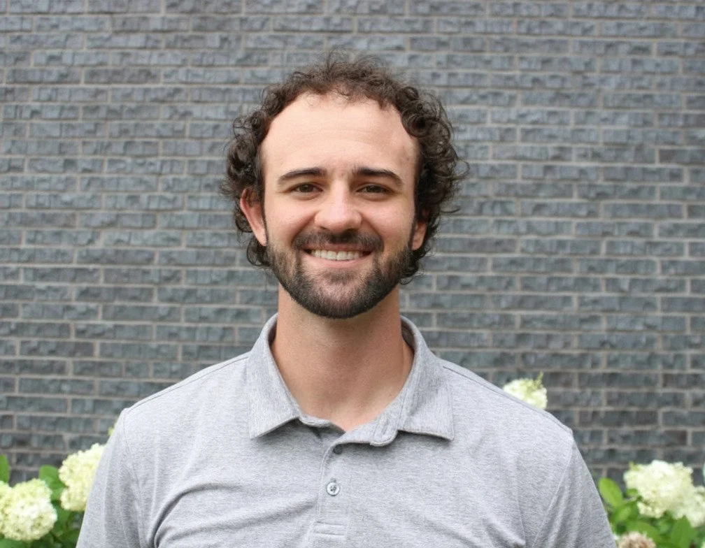 Smiling person in blue plaid shirt standing in front of a brick wall with dried flowers.