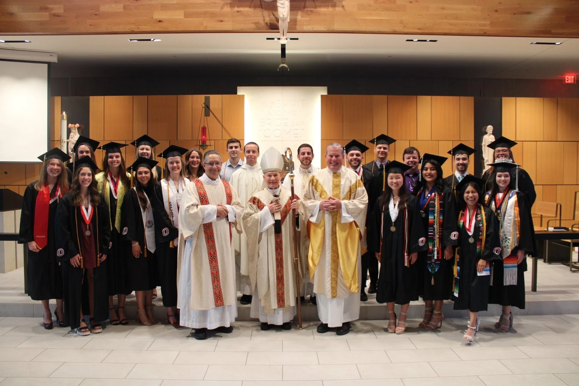 Group of graduates in caps and gowns with clergy in a church setting.