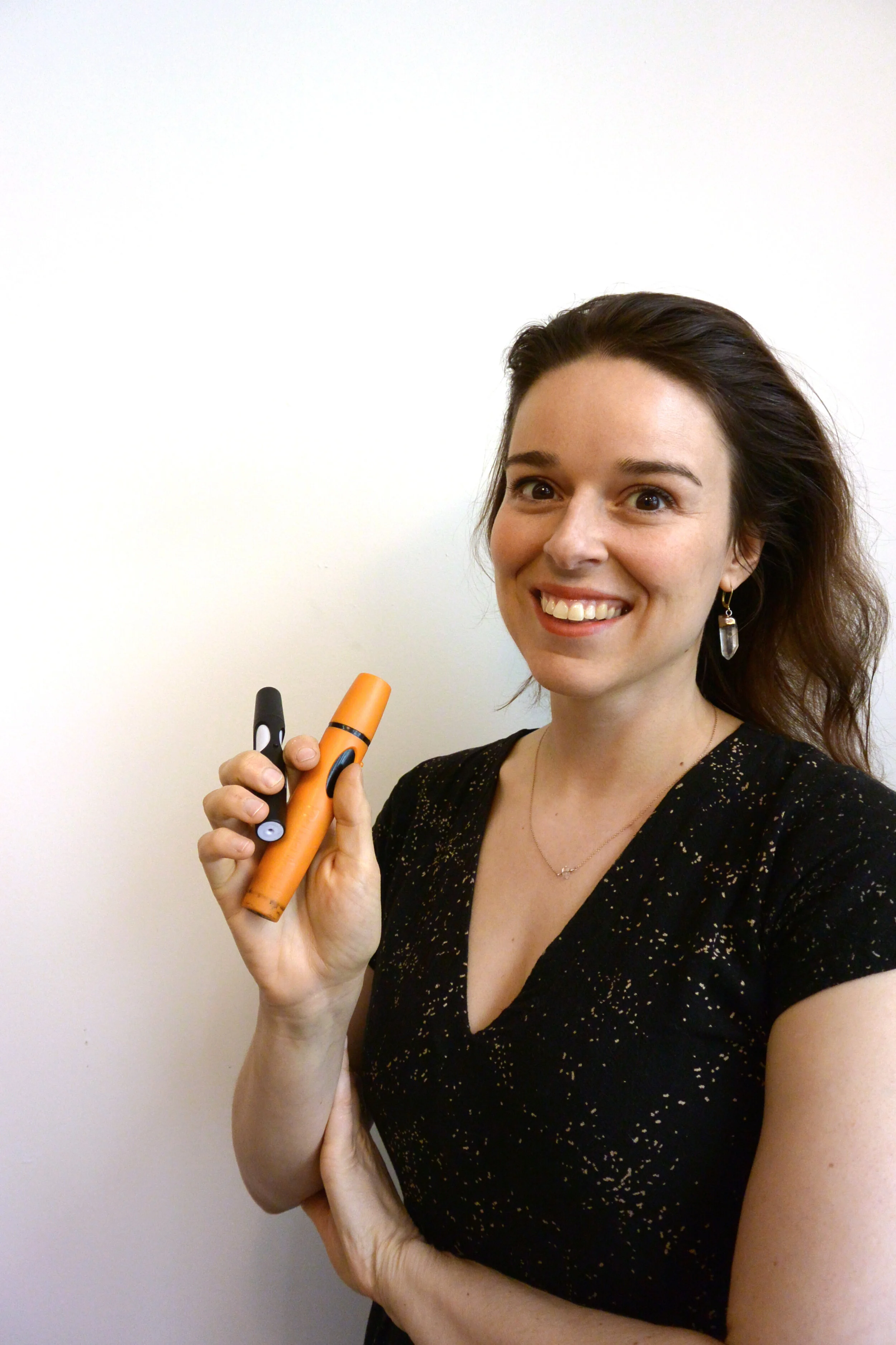 A woman with long brown hair smiling while holding two markers against a white wall