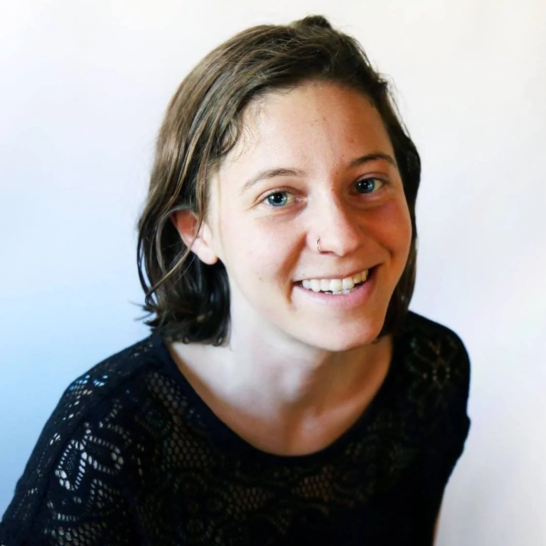 A young woman with short brown hair, blue eyes, and a nose ring smiling at the camera, wearing a black lace top, against a plain white background.