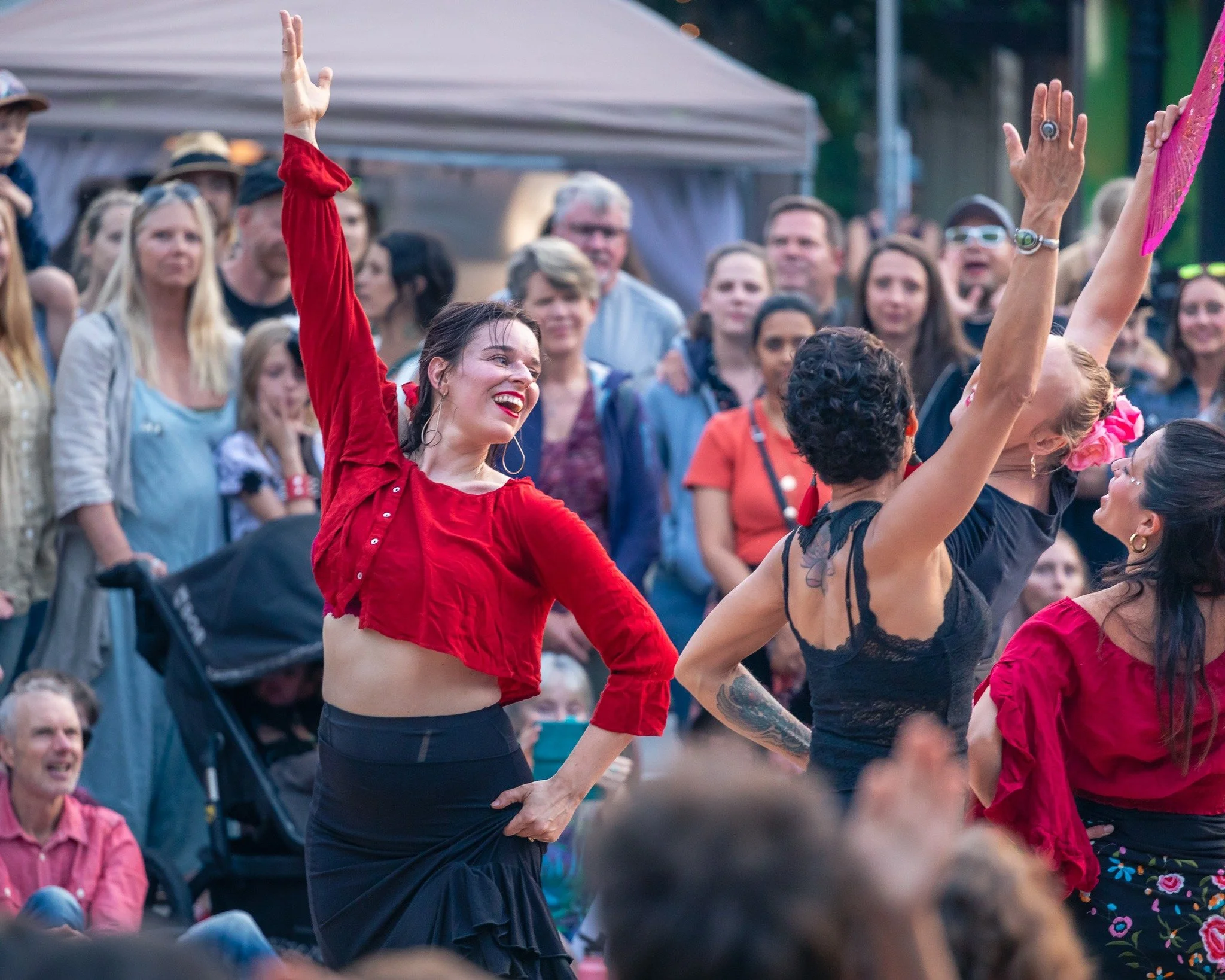 A woman in a red crop top and black skirt dancing flamenco with a smile at an outdoor event, surrounded by a crowd.