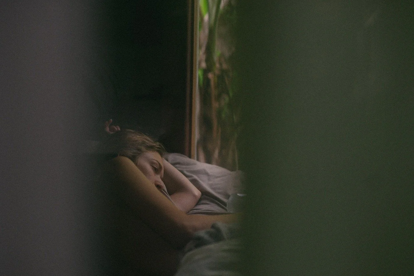 A young woman sleeping on her side with her head resting on her arm, covered by a blanket, in a dimly lit room with a window showing trees outside.