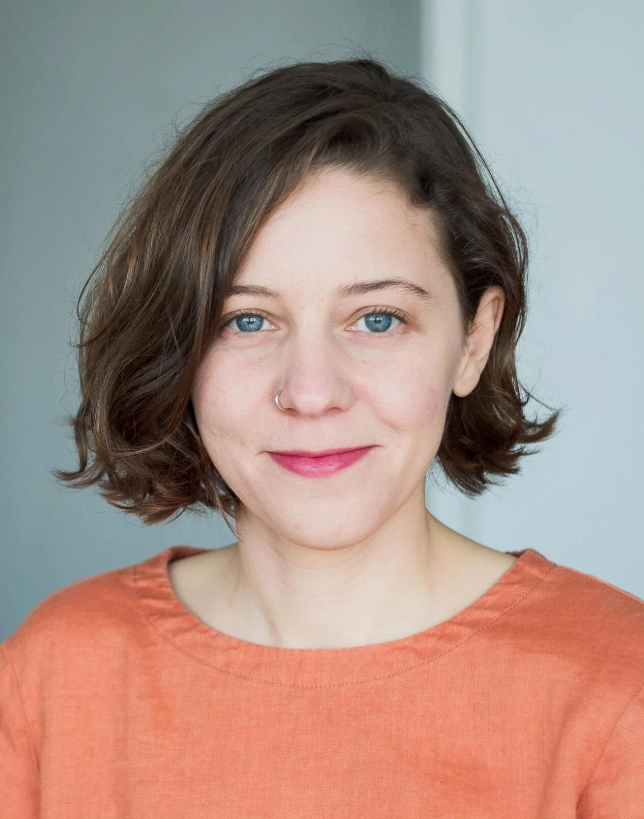 A woman with shoulder-length dark brown hair and blue eyes, wearing a light peach-colored top and a small nose ring, smiling softly at the camera against a neutral background.