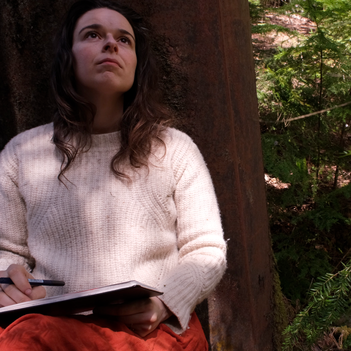 Woman with dark wavy hair wearing a white sweater, sitting outdoors near greenery, with a rusty metal structure behind her, holding a notebook and pen, looking thoughtfully upward.