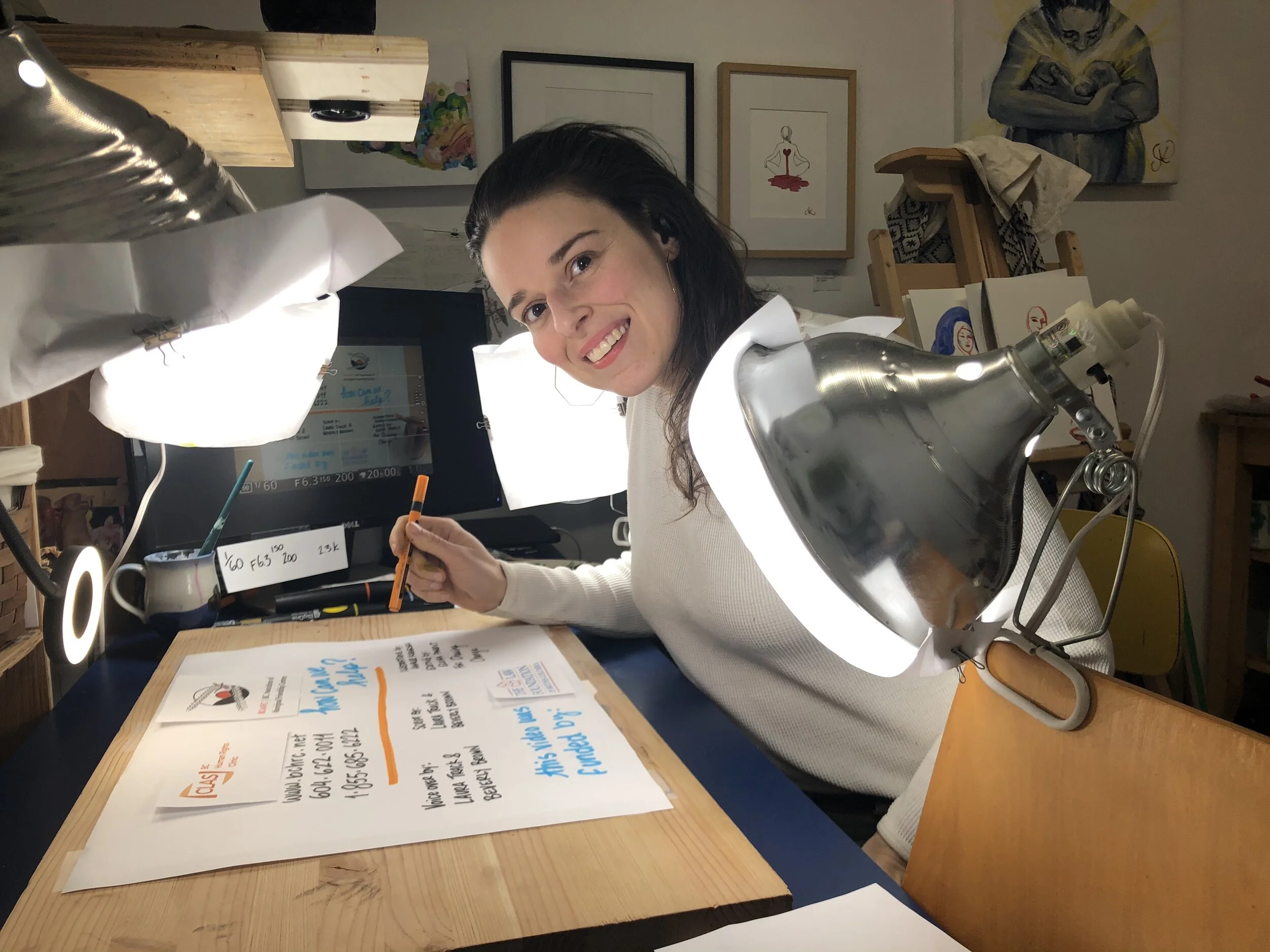 A woman smiling and working at a desk illuminated by bright lights. She is drawing a hand-animated video. There are artwork and picture frames hanging on the wall behind her.