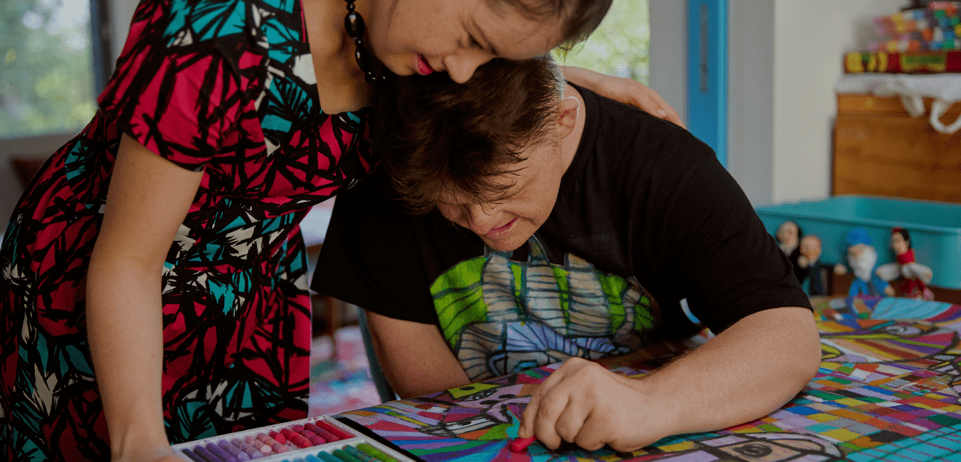 Two friends wearing colourful clothes create a colourful artwork on a table, surrounded by art supplies and plush toys in the background.