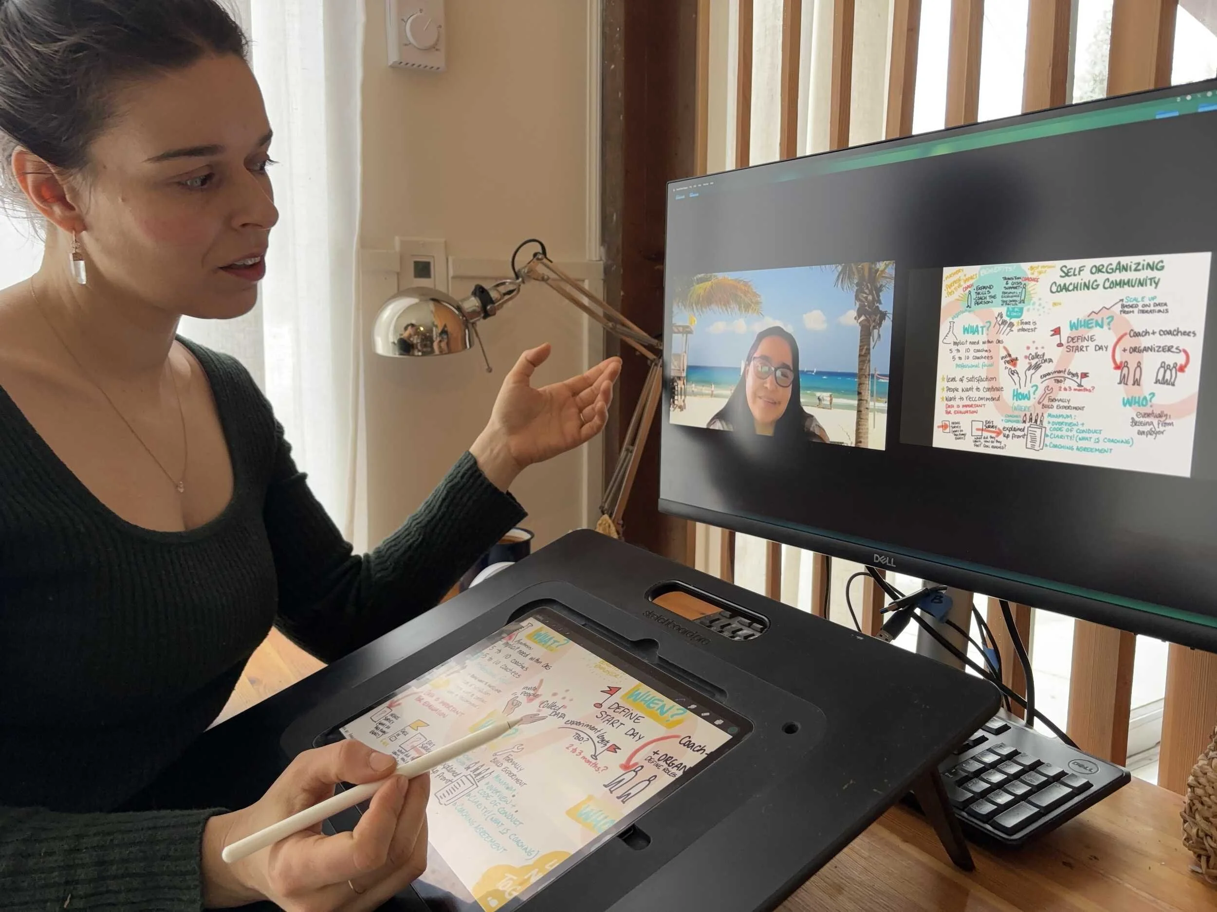 Woman sitting at a desk using a digital drawing tablet with a stylus, participating in a video conference on a computer monitor. The monitor displays a woman's face on a beach background and a colorful infographic about coaching community. The room has wooden window blinds and a desk lamp.