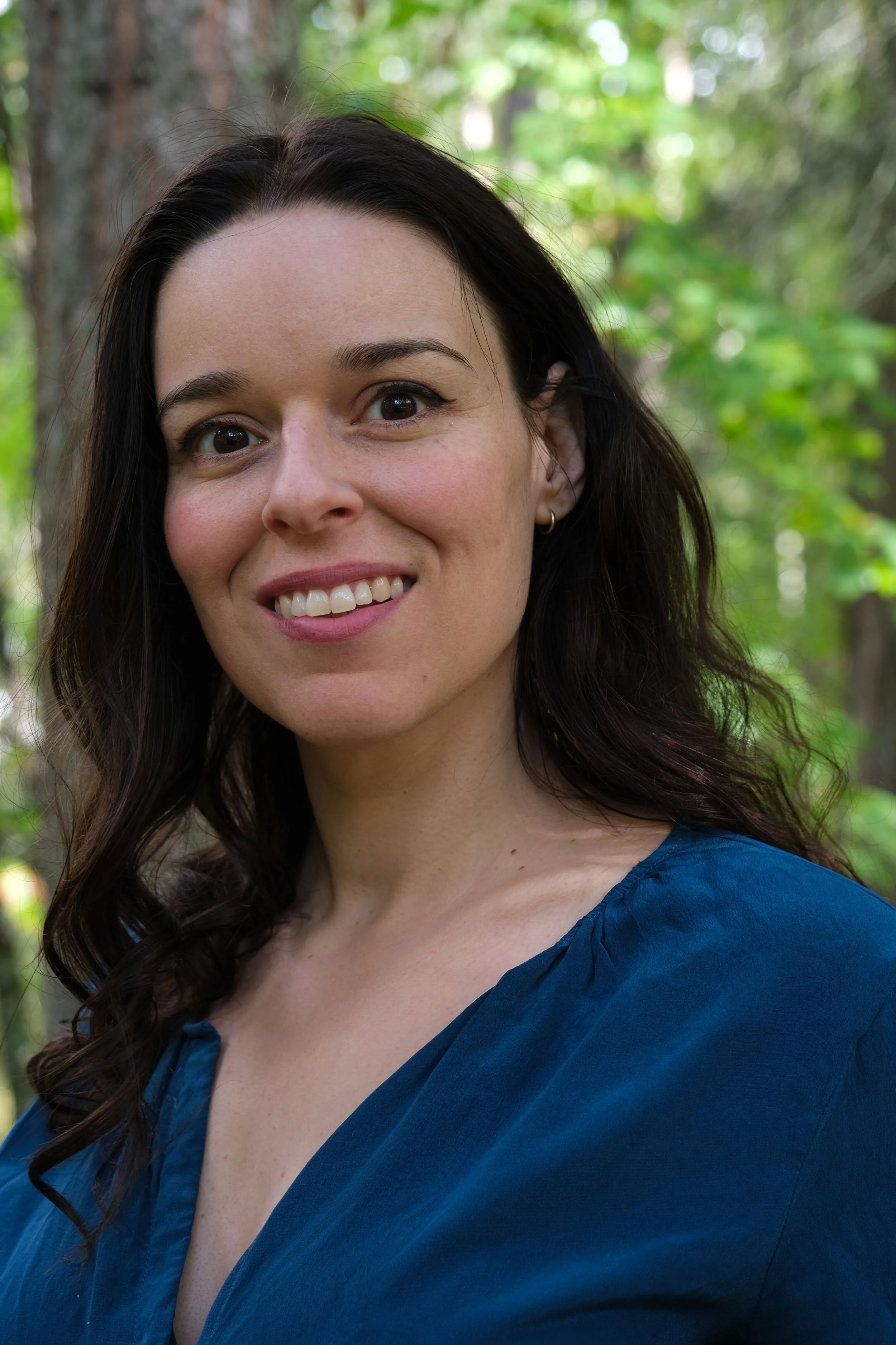 A woman with dark brown hair, smiling, standing outdoors in a forested area wearing a blue top.