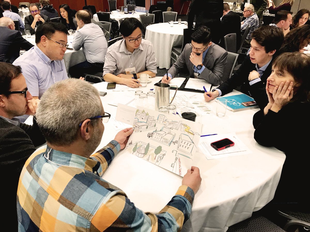 Group of people sitting around a round table, studying a large illustrated map, in a conference or meeting room setting.