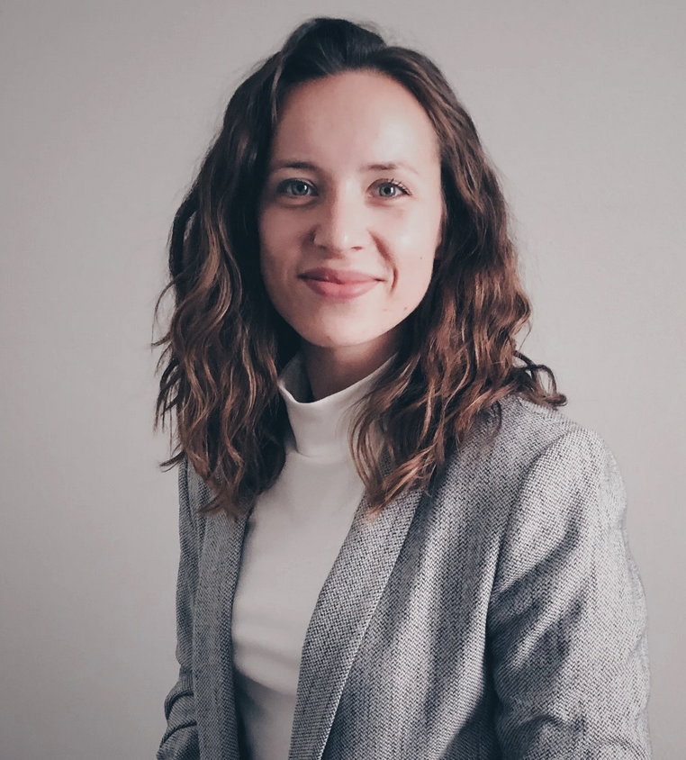 A woman with short dark hair sitting on a wooden stool in front of a white wall with a beige leafy pattern. She is wearing a black V-neck top and dark jeans, and has earrings. She is looking at the camera with a slight smile.