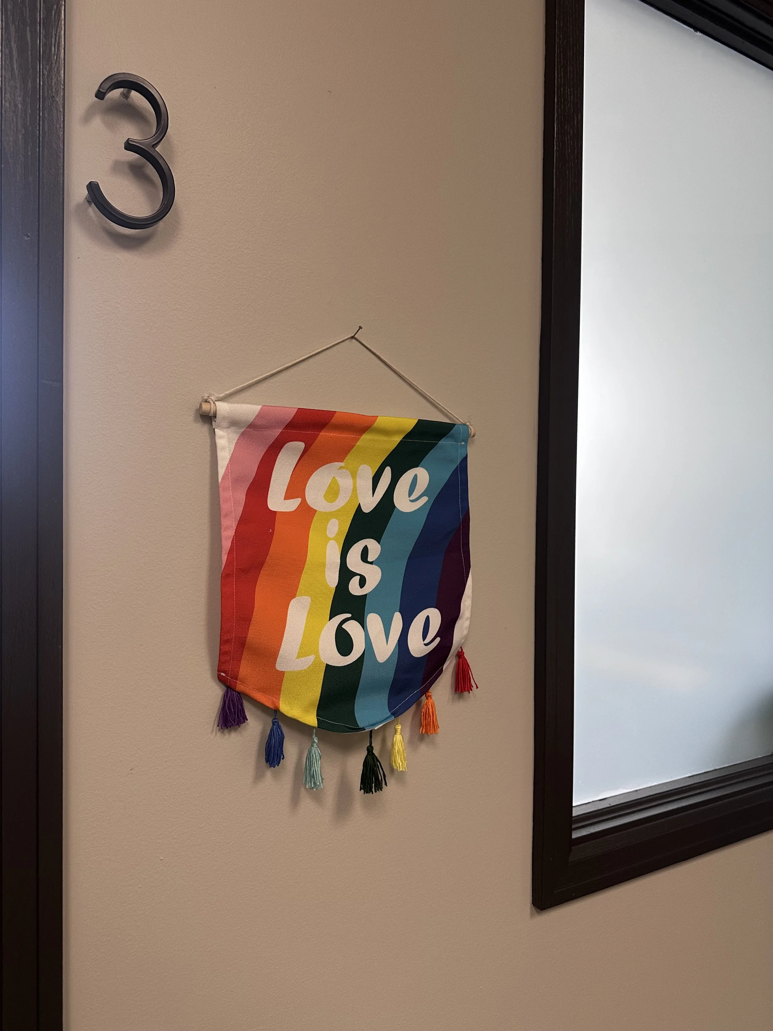 A rainbow-colored flag with the words 'Love is Love' hanging on a wall, with colorful tassels at the bottom.