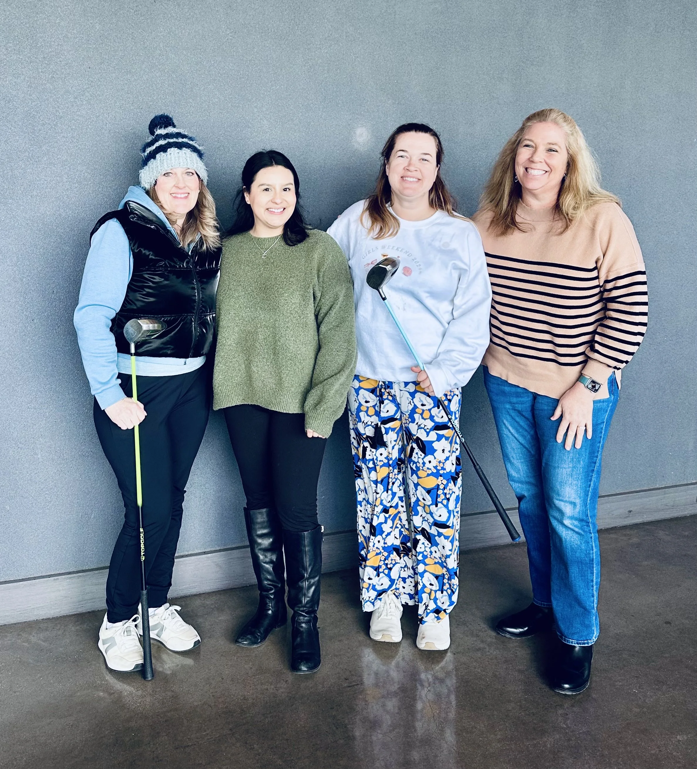 Four women standing against a gray wall at a golf course, smiling. The woman on the far left wears a winter hat, black vest, blue hoodie, and white sneakers, holding a golf club. The second woman is in a green sweater, black pants, and black boots. The third woman wears a white sweatshirt with a golf-themed design, colorful patterned pants, and white sneakers, holding a golf club. The woman on the far right has long blonde hair, wears a tan and black striped sweater, blue jeans, and black boots.