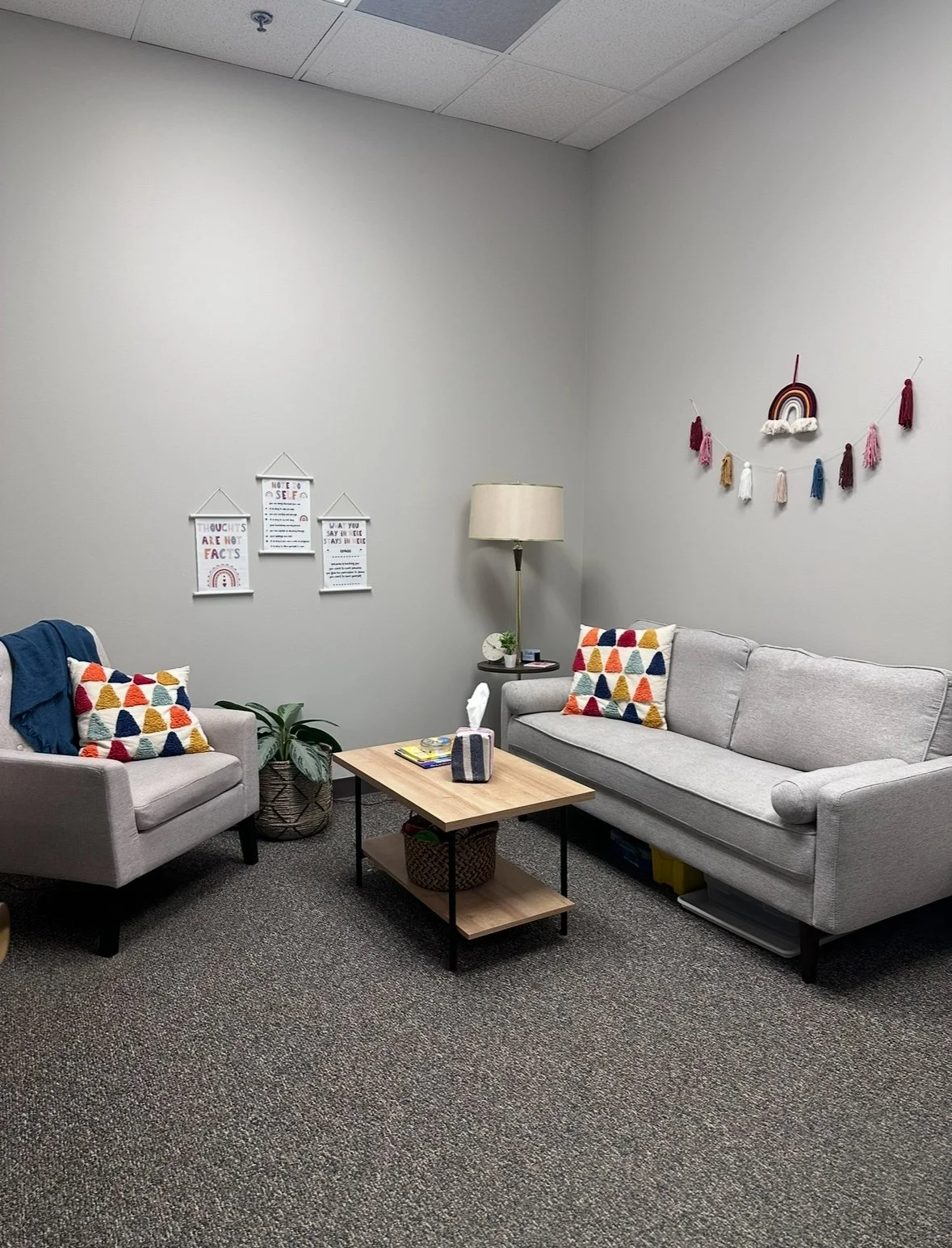A cozy office or waiting room space with two light gray sofas, colorful triangular-patterned pillows, a wooden coffee table with tissue box and magazines, a tall floor lamp, and wall decorations including wall hangings and framed quotes.