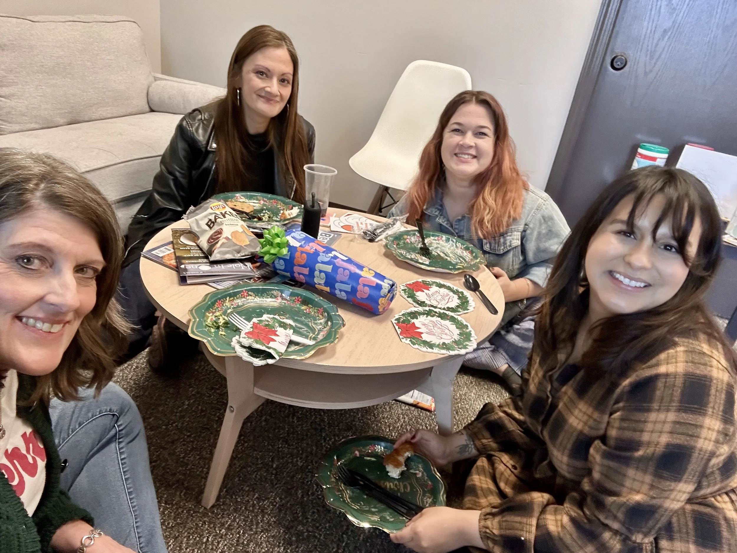 A group of women gathered around a table at a holiday party, smiling. The table has Christmas-themed plates and snacks, with a woman in a plaid shirt holding a plate and fork.